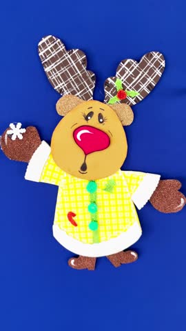 Vertical 4K video of a handmade DIY Christmas reindeer craft made from colorful foam sheets. Cute holiday decoration with plaid antlers and red nose isolated on a blue background.