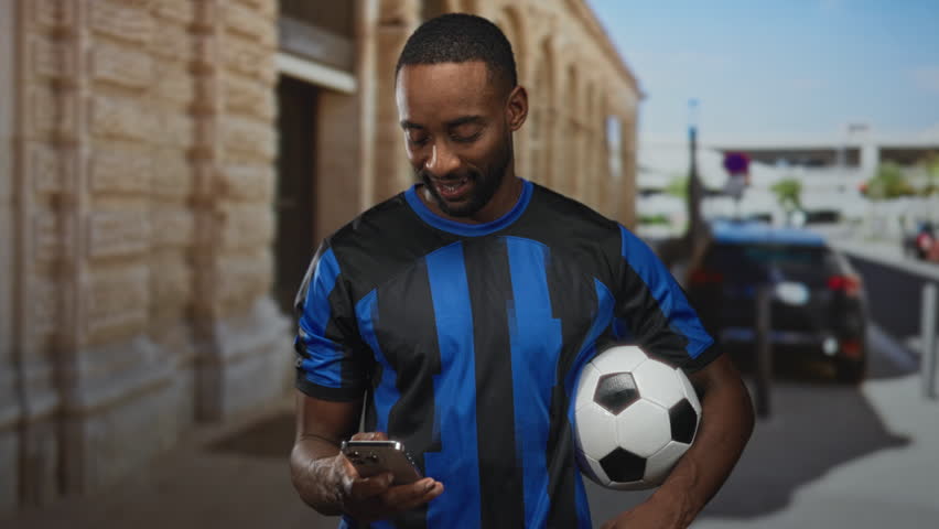 Man holds soccer ball under arm and checks smartphone on street by building entrance; concentration team spirit.