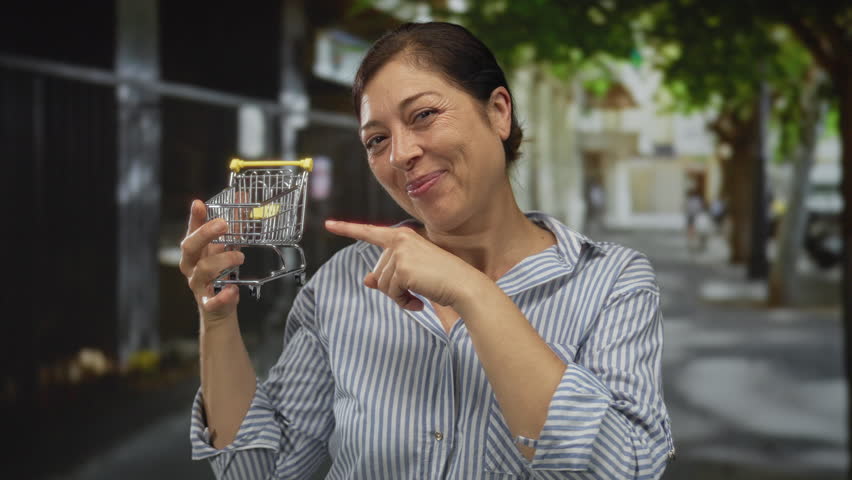 Woman holding miniature shopping cart smiles and points finger to tiny cart on sunlit street; shopping joy.