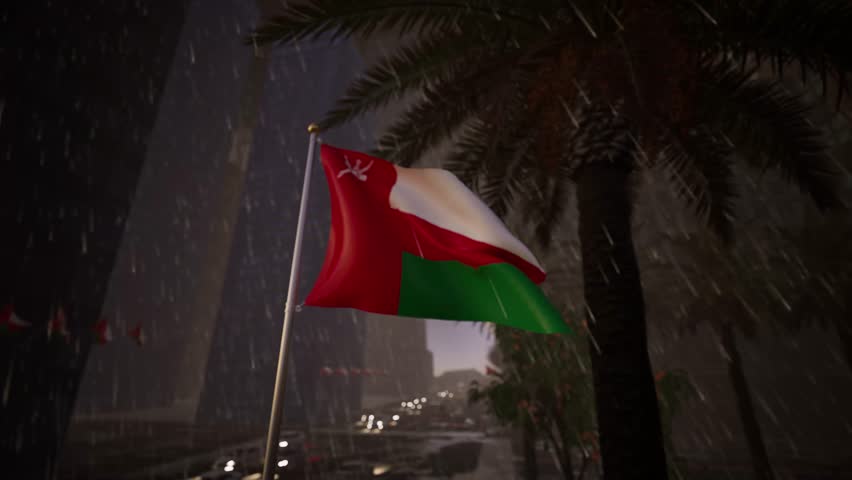 Oman national flag waving in heavy rainfall across a modern city street with wet reflective surfaces, palm trees and moving traffic. This dramatic scene likely depicts the unique Khareef monsoon season in the southern region of Dhofar, specifically Salalah, or a rare winter storm in the north. The fabric is heavy with moisture, struggling against the gusts of wind and the weight of the water, yet it remains aloft, symbolizing resilience against the elements. Raindrops streak through the frame, blurring the background of lush greenery and wet asphalt. The streetlights reflect on the slick road, creating a mirror-like effect that duplicates the colors of the ensign and the passing vehicles. The usually arid environment is transformed into a wet landscape, showcasing the diverse climate of the Sultanate. The sound of the downpour and the whipping of the wet textile adds to the intensity. This clip is invaluable for meteorology reports, climate change discussions in the Middle East, or travel features about the green season in Arabia. The colors of the banner appear deeper and more saturated due to the dampness. It portrays a side of the Gulf often unseen by the outside world, breaking the stereotype of perpetual desert dryness. The pendant flutters erratically, shedding water with every snap.