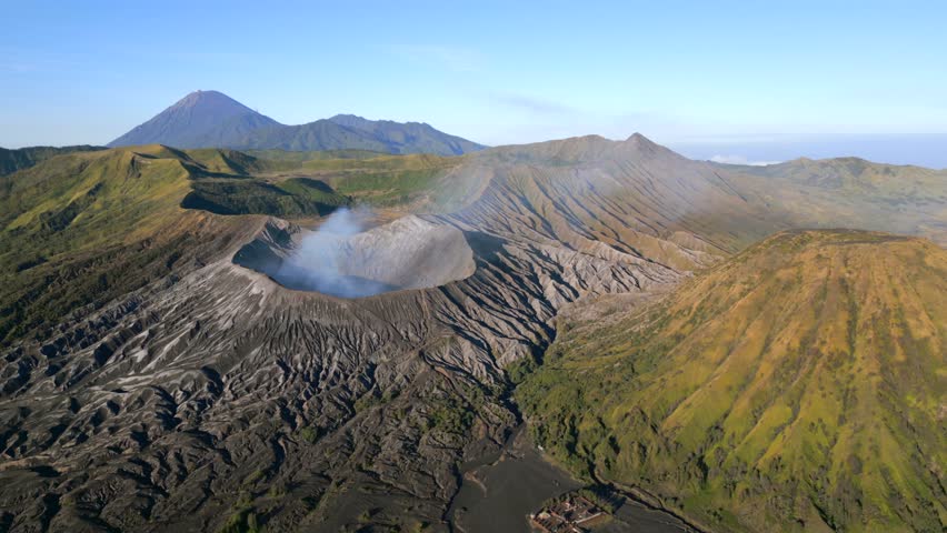 Aerial view of active Mount Bromo volcano emitting smoke with Mount Semeru in the distance on sunny day. Dramatic volcanic landscape in East Java, Indonesia.