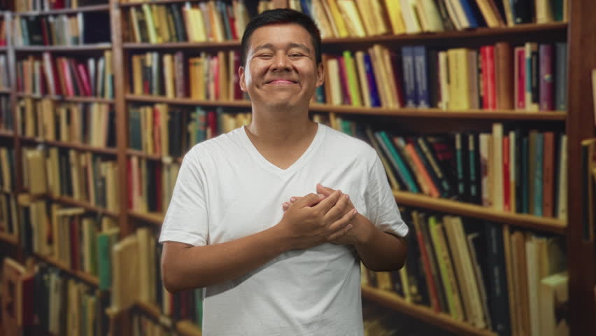 Hispanic man wearing white t shirt hands on chest in library building smiling with closed eyes; gratitude reflection.