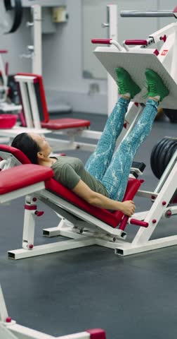 Gym workout. Vertical 4k footage of a middle-aged woman trains her leg muscles with the leg press machine, focusing on controlled, powerful movements during her gym workout.