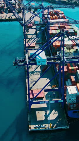 Container ship docked in sea port with cargo operation containers by means of gantry cranes in port. Vertical drone video