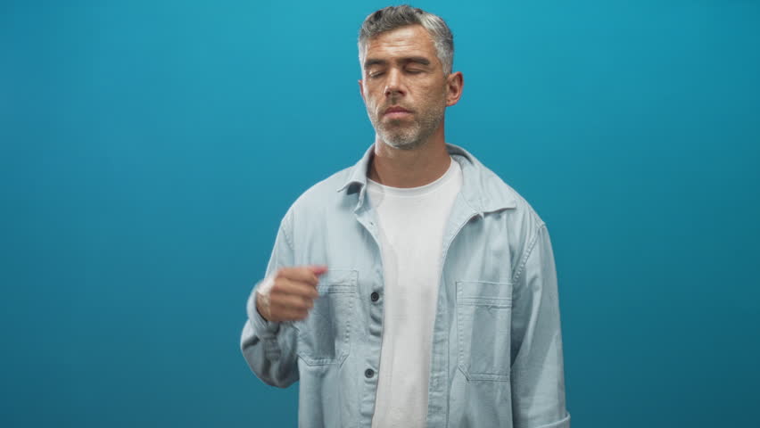 Man with outstretched left hand palm up presenting an empty palm, smiling and looking at his hand while wearing a light denim jacket against a studio teal backdrop; calm invitation.