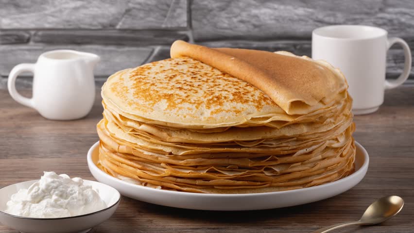 Stack of golden crepes served with sour cream on a wooden table