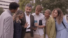 Group of multi ethnic business colleagues standing outdoors, looking at a tablet. Senior manager explains a project to his diverse team, who are listening and smiling - Powered by Shutterstock - Get 15% off with code: PIKWIZARD15