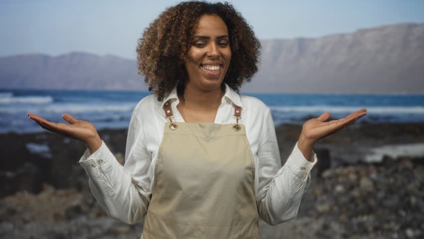 Young african american woman baker in apron smiling with open palms on rocky seaside coast outdoors; joy baking community.