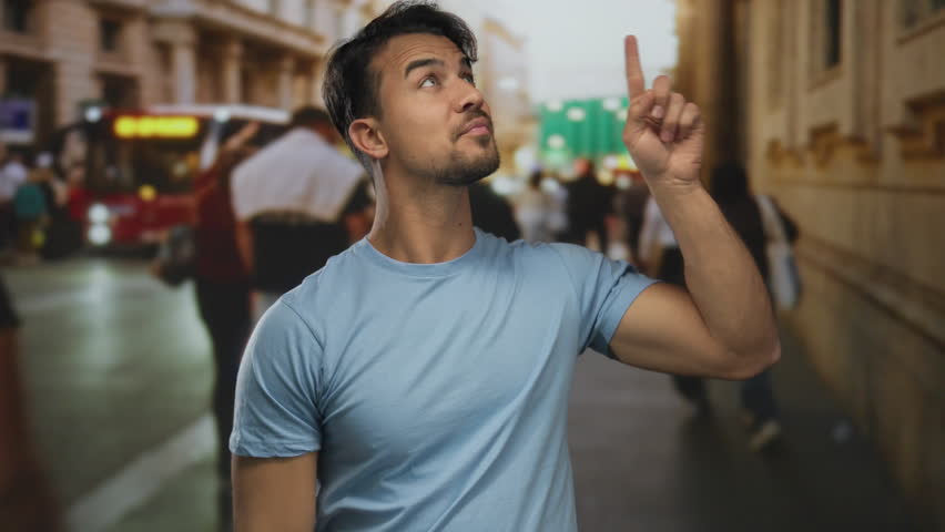 Young hispanic man smiling and giving a thumbs-up on a bustling city street with blurred people and buildings in the background.