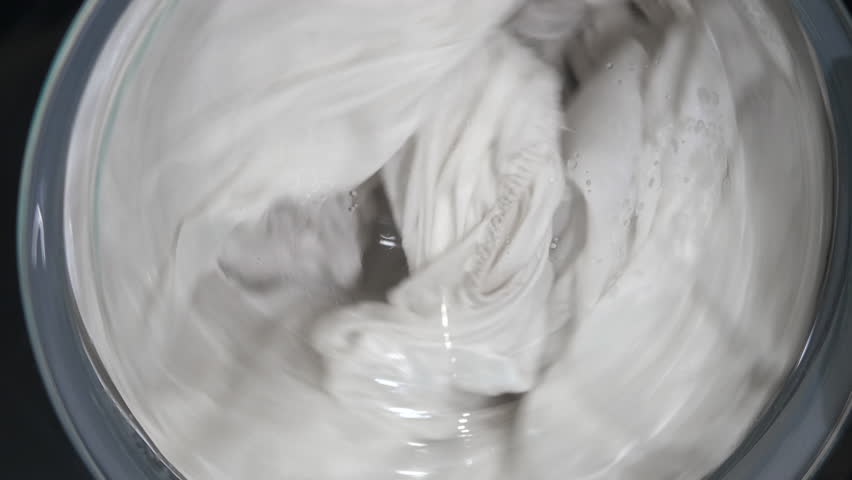 White laundry washing machine cycle spinning. Close-up top view of white clothes spinning inside a washing machine drum during a wash cycle, showcasing the rotating motion of the laundry with water