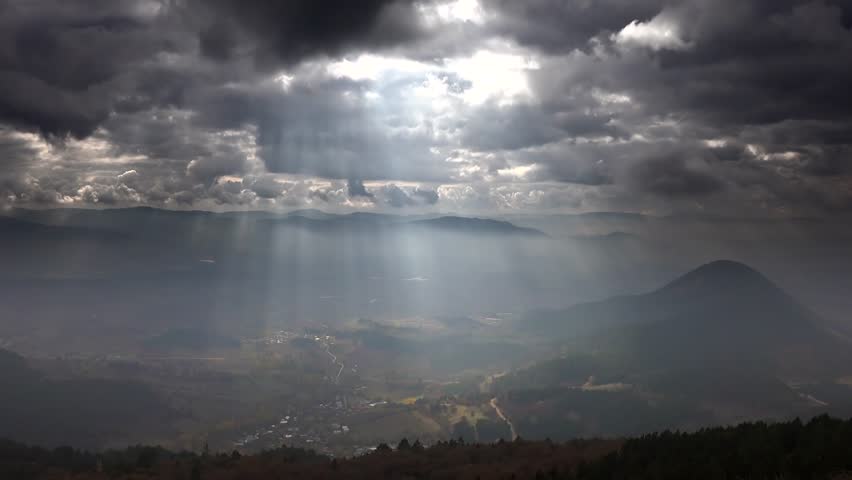 Time lapse view from mountain peak as sun rays break through clouds above a cone hill on the plain. Atmospheric beams pierce overcast skies and cast soft shifting light across the wide plain below.