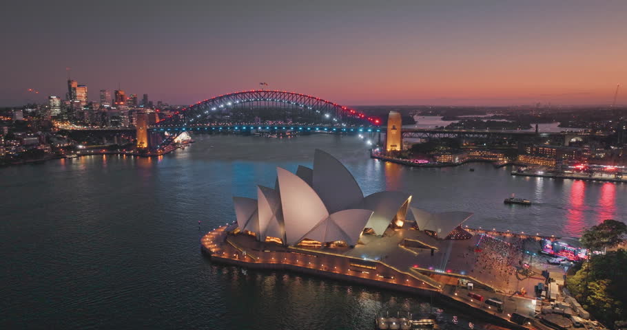 Sydney Opera House with Sydney Harbour Bridge in the background illuminating night lights, creating a breathtaking cityscape view with colorful sky and illuminated buildings. Aerial view drone footage