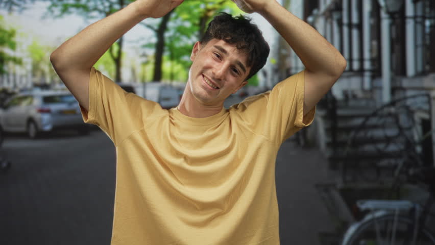 Man raises hands above head on a busy city street, smiling with closed eyes and visible forearms while standing near parked bicycles; youth optimism joy.