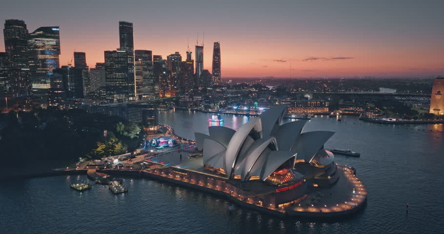Australia, New South Wales: Night colorful Sydney Opera House and skyline reflecting on water during sunset, colorful sky and illuminated city lights beautiful cityscape. Aerial view drone footage