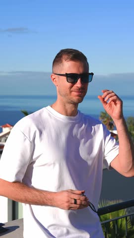 A man in a white T shirt stands on a terrace by a metal railing, adjusting sunglasses, with a second pair in hand, Funchal coast and Atlantic behind in warm light.