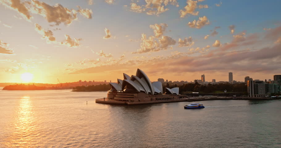 Australia, Sydney Opera House at sunset, bright warm orange sunlight over the harbor, passenger ferry crosses calm water with the city skyline in background. Travel destination. Aerial drone panorama