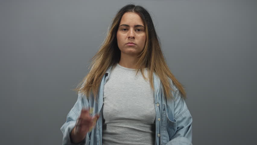 Woman standing with hand on cheek and folded arm wearing denim shirt in gray studio; pensive fatigue reflection.