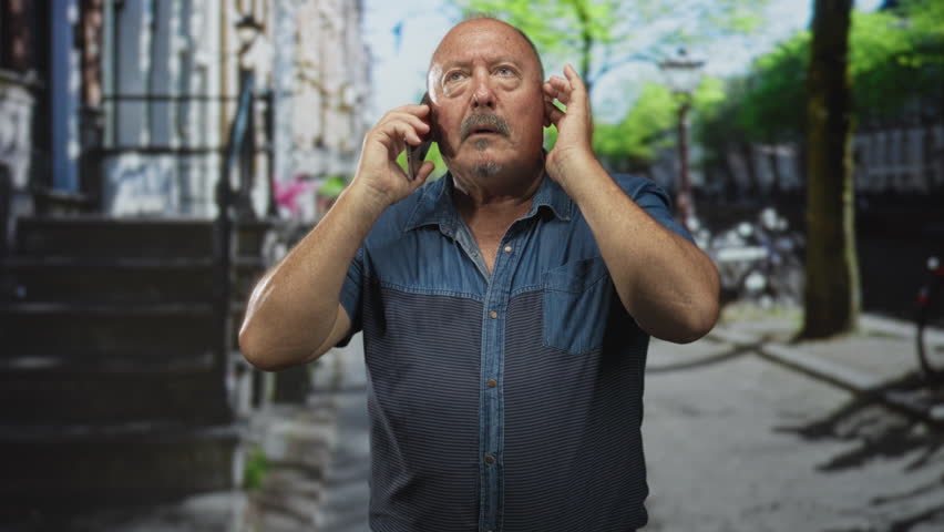 Man in blue denim shirt holds smartphone to his ear and cups the other ear while standing on a tree lined street with parked bicycles; concern.