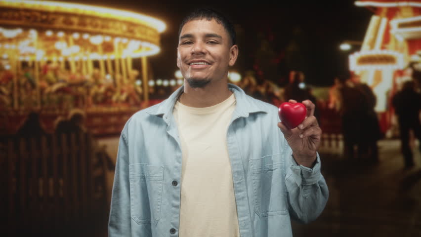 Man holding red heart and smiling, wearing light jacket, looking sideways at bright carnival lights on a street with carousel and blurred crowd; love hopeful.