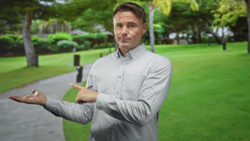 Handsome man points finger to palm showing lotion on hand in forest path near trees and walkway; product demo confidence.