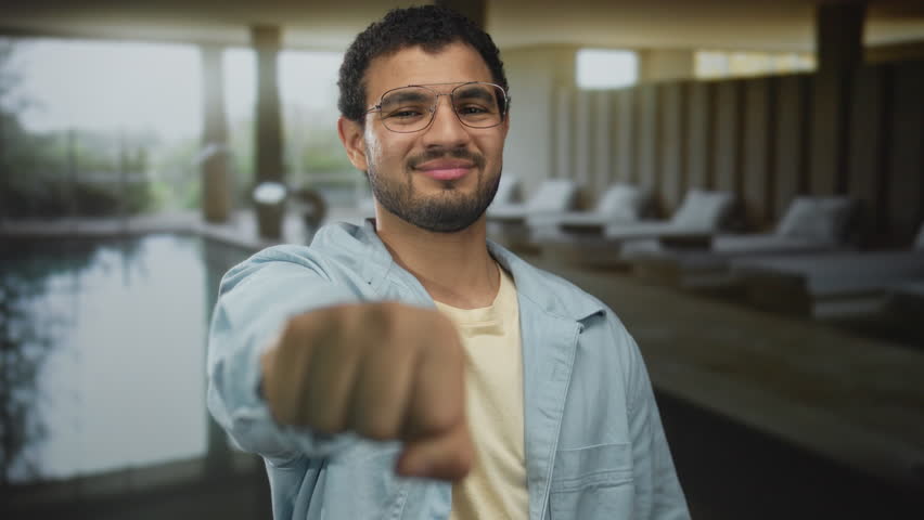 Hispanic man wearing glasses smiles and extends fist toward camera in indoor spa building; confidence.