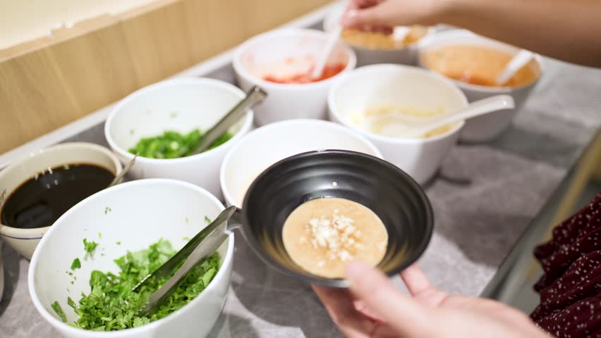 Hand adds chili to sesame sauce at a buffet condiment station, bright indoor lighting, close-up