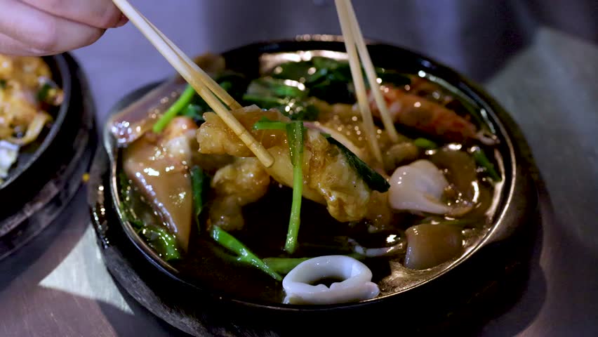 Chopsticks lift seafood pad thai noodles from a sizzling hot plate under night lighting.