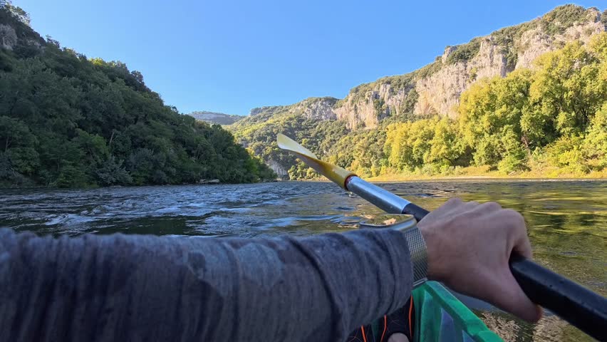 First-person view paddling a green kayak with a yellow paddle down a scenic river in Auvergne, France. Rocky cliffs and lush green forests line the banks on a beautiful sunny summer day