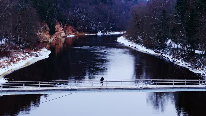 Frozen Gauja flows beneath woman dancing in solitude on pedestrian bridge