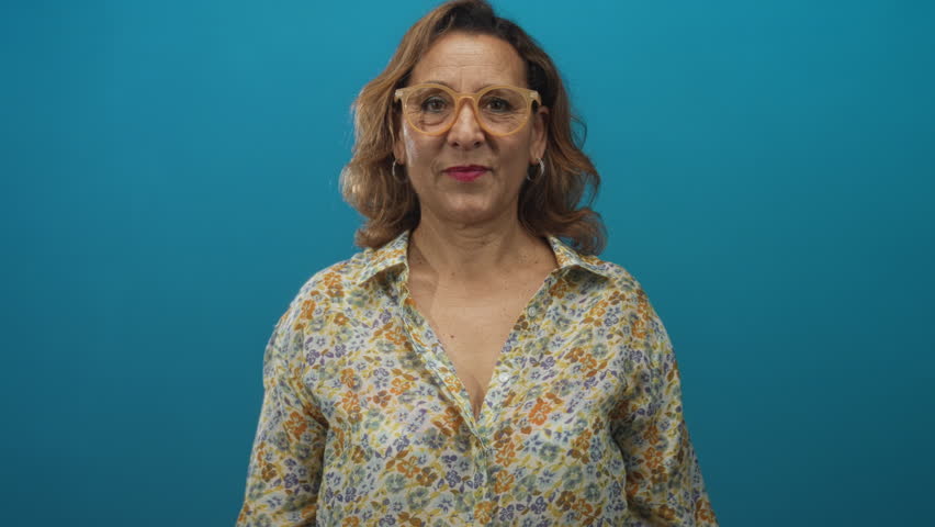 Woman wearing glasses and floral blouse pointing thumbs at herself in blue studio; confidence empowerment.