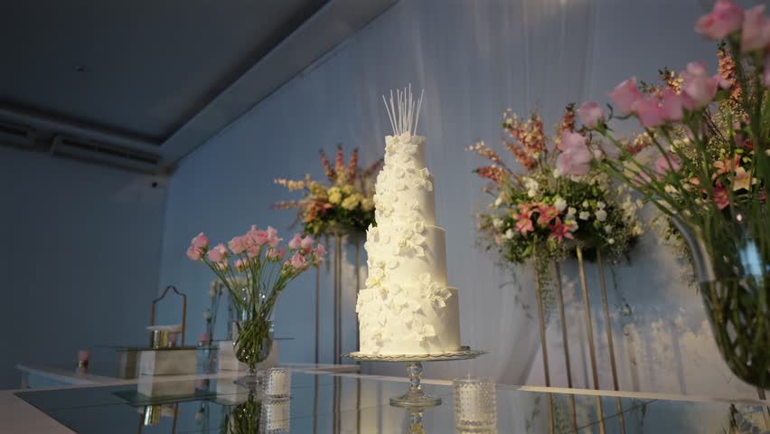 Wide shot of a beautiful, classic multi-tiered wedding cake decorated with flowers and set among pink floral arrangements.
