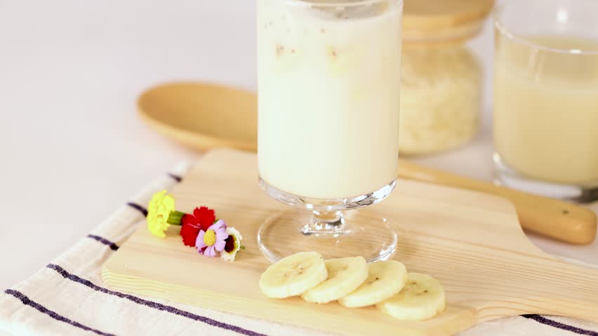 Hand stirs banana milkshake in glass with striped straw, bright lighting, minimal kitchen background