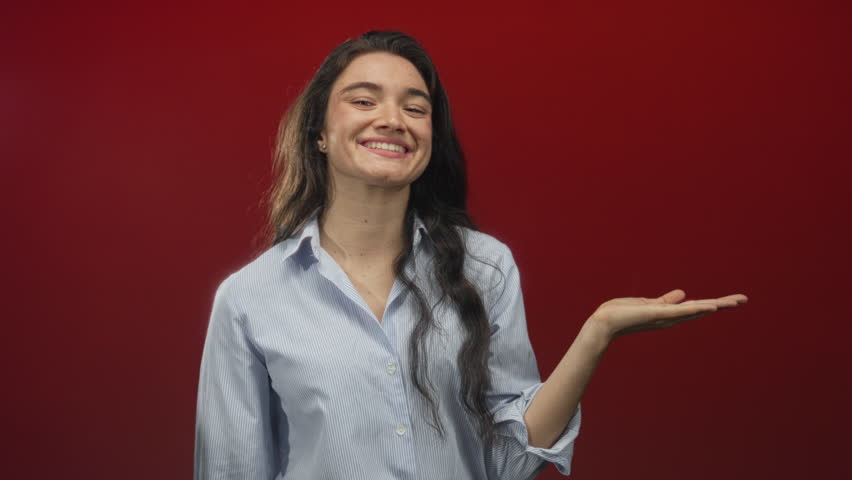 Hispanic teen woman in striped shirt pointing finger to open palm in red studio setting; confidence.