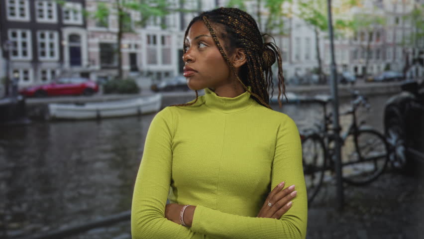 Woman in green sweater with arms crossed, ring and bracelet visible, standing by canal on an amsterdam street with bicycles and canal houses; quiet confidence.