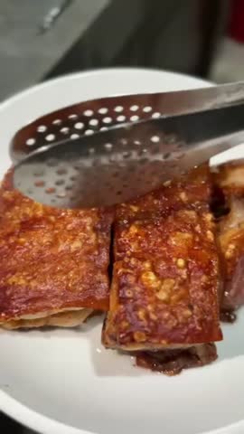 Extreme close-up of crispy, golden-brown roasted pork belly slices being handled with tongs on a white plate. Focus on the crunchy, crackling skin.