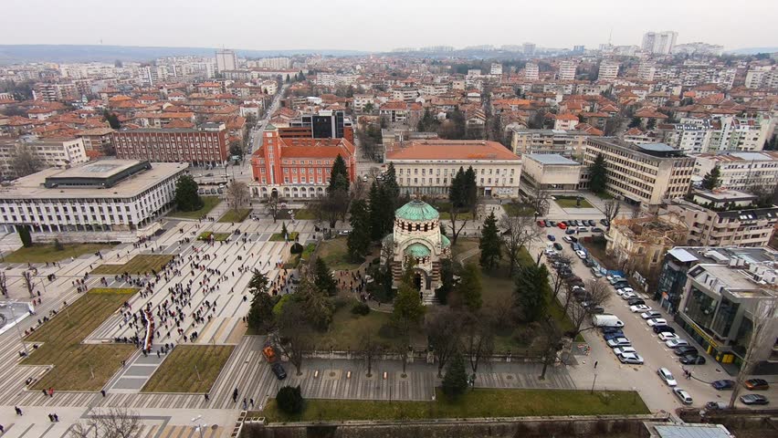 Drone video of Pleven center on March 3, 2019, showing landmarks, the square, the Ossuary, church, municipal building, fountains, and people walking with a long national flag, Pleven, BG,03 March 2019