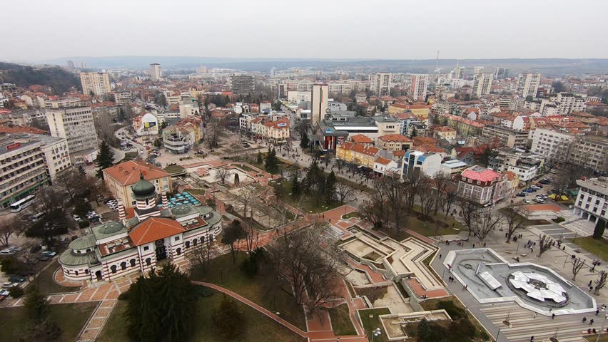 Drone video of Pleven center on March 3, 2019, showing landmarks, the square, the Ossuary, church, municipal building, fountains, and people walking with a long national flag, Pleven, BG,03 March 2019