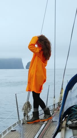 A Voyager in an Orange Raincoat Stands Gracefully on a Sailboat, Observing the Calm Sea and Misty Landscape, Embracing the Serenity of Nature's Embrace