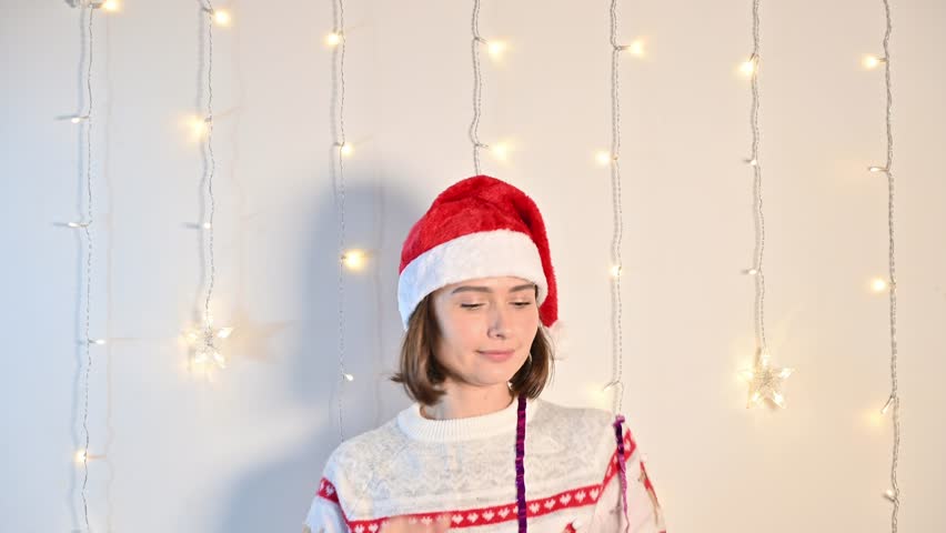 A young woman wearing a festive Christmas sweater and Santa hat joyfully dancing on a white background with decorative lights, celebrating the holiday season.