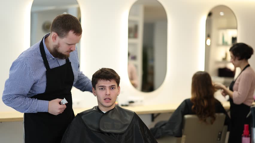 Male hairdresser stylist gives haircut to young guy client in beauty salon