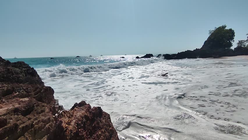 Scenic coastal view with waves crashing onto rocky shores under bright sunlight, capturing the serene beauty of a clear blue sea and natural beach landscape