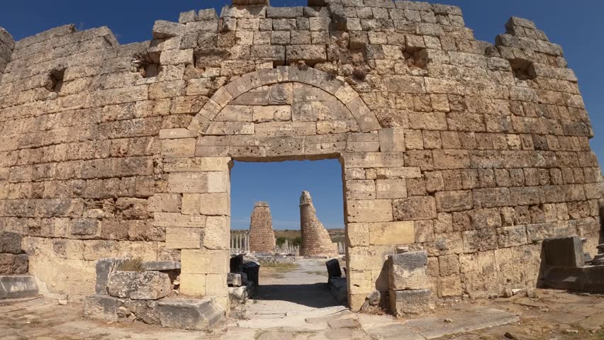 Aerial view of ancient stone archway leading toward twin pillars within historic Patara ruins in Antalya Province, Turkey, surrounded by weathered masonry and open landscape.