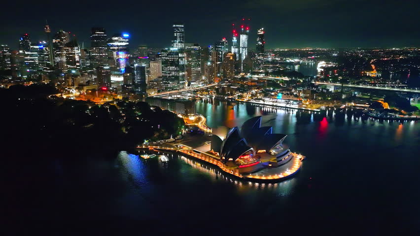 Australia, New South Wales, Sydney: Bright colorful lights illuminate Sydney Opera House building and city business center skyline skyscrapers glowing at night. Aerial view drone footage panorama