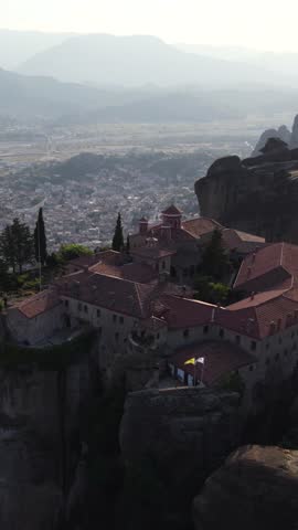Vertical aerial reveal of Agios Stefanos Monastery on a precipice above Kalabaka, capturing the rocky terrain and town scenery.