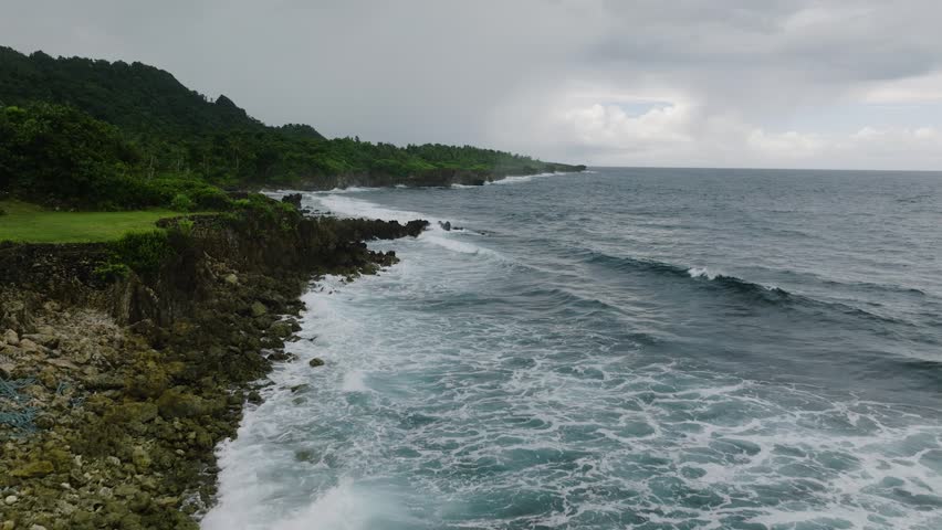 Ocean waves crash against rugged rocky cliff with lush vegetation under cloudy sky. Siargao, Philippines.