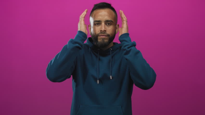 Man pressing hands to temples with eyes closed in magenta studio, wearing teal hoodie and rings on fingers, head bowed slightly; frustration.