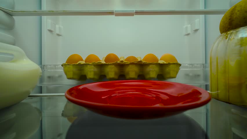 Man opening fridge, indifferently looking inside and chewing, view from inside of fridge. Male adult feeling bored and distracted. Guy eating something while checking fridge content