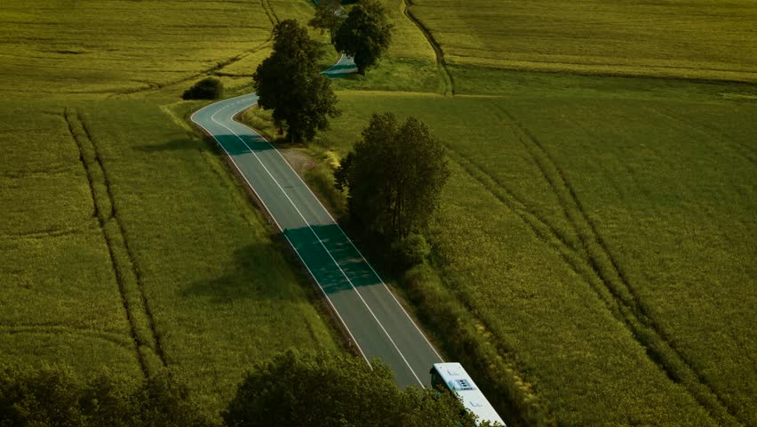 Transport driving through curved road in open fields at sunset. White bus moving along countryside highway under dry sunny weather. Vehicle traveling past farmland during golden hour