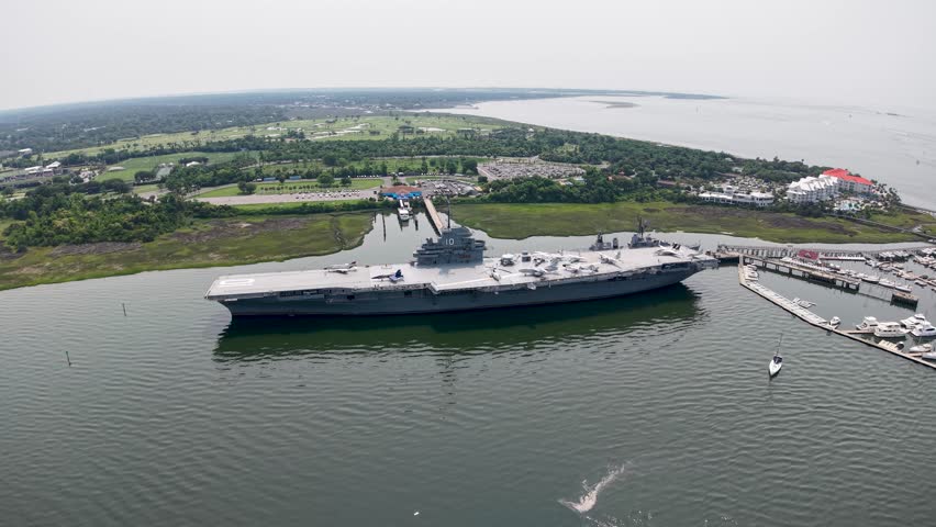 Aerial drone clip sweeping over the USS Yorktown aircraft carrier docked at Patriots Point in Charleston, with nearby pier, boats and surrounding marshland visible under clear skies.