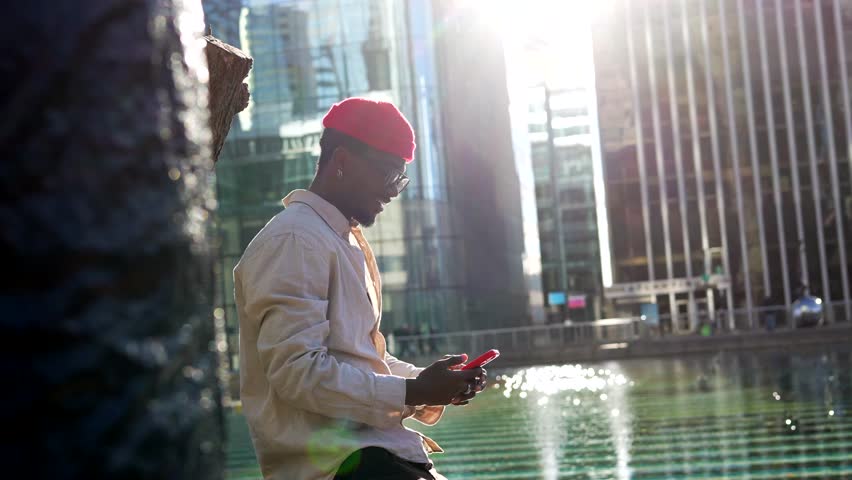Happy young black man in a red beanie using his smartphone for a video call, smiling and gesturing while sitting by a fountain with modern skyscrapers and bright sunlight in the background